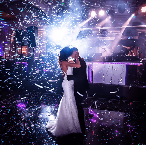 Bride and groom embracing and kissing on a dance floor covered with confetti under colorful lights at a wedding reception.