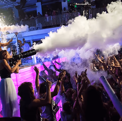 Woman using a smoke cannon to release dense white smoke over an excited crowd at a vibrant indoor party.