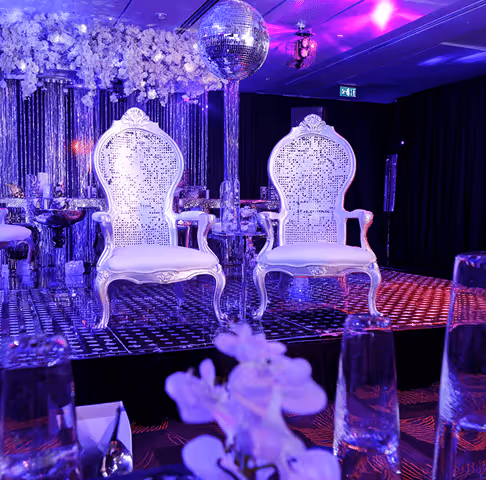 Two ornate white chairs on a stage under purple lighting with a disco ball overhead and table with flowers and glasses in the foreground.