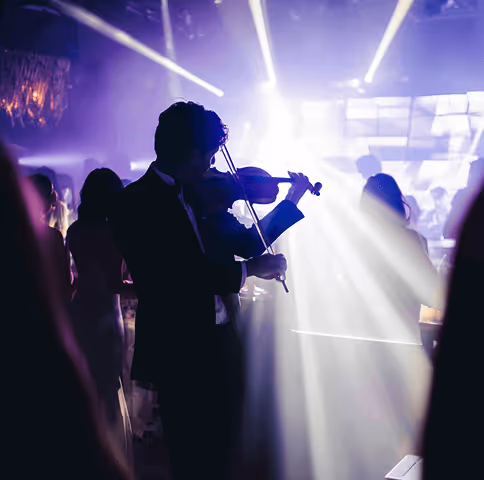 Silhouette of a man playing violin amid bright stage lights and an audience in a dimly lit venue.