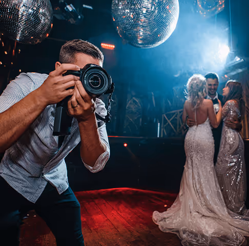 Photographer taking a picture at a wedding with a bride in a white dress and guests under disco balls in a dimly lit venue.