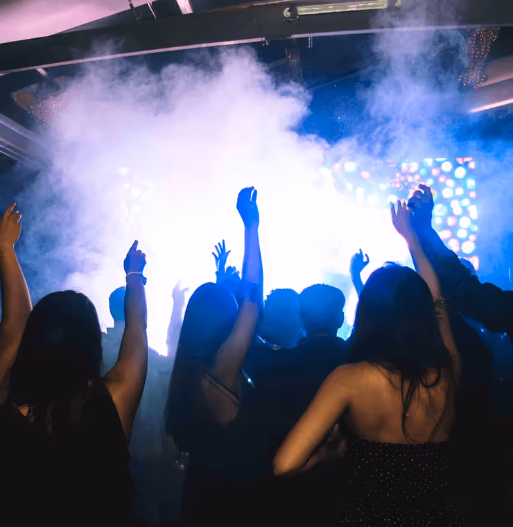 People at a concert or club with hands raised amid bright lights and smoke effects.