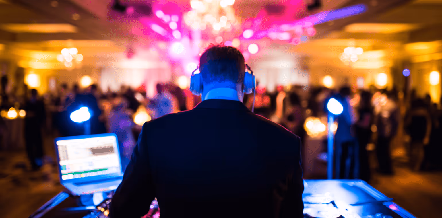 DJ wearing headphones working at a laptop and mixing console in a dimly lit party venue with colorful lights and dancing crowd.