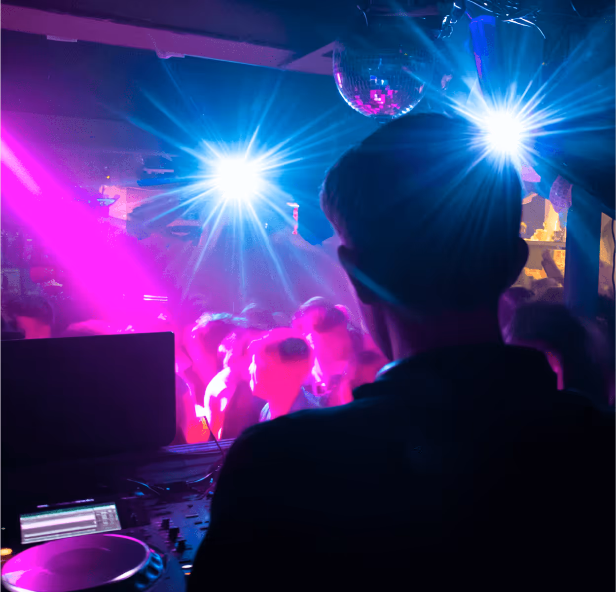 DJ performing in a dimly lit club with colorful purple and blue lights and a crowd dancing in the background.