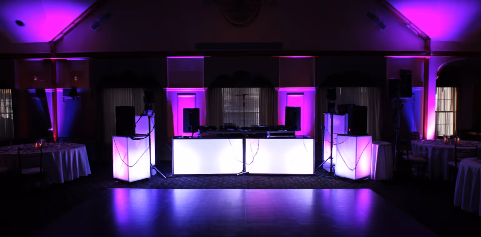 Dark event space with DJ booth illuminated by purple and white lights and empty dance floor.