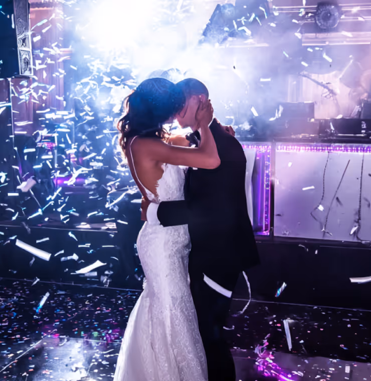Bride and groom kissing on dance floor amidst falling confetti and colorful lights at wedding reception.