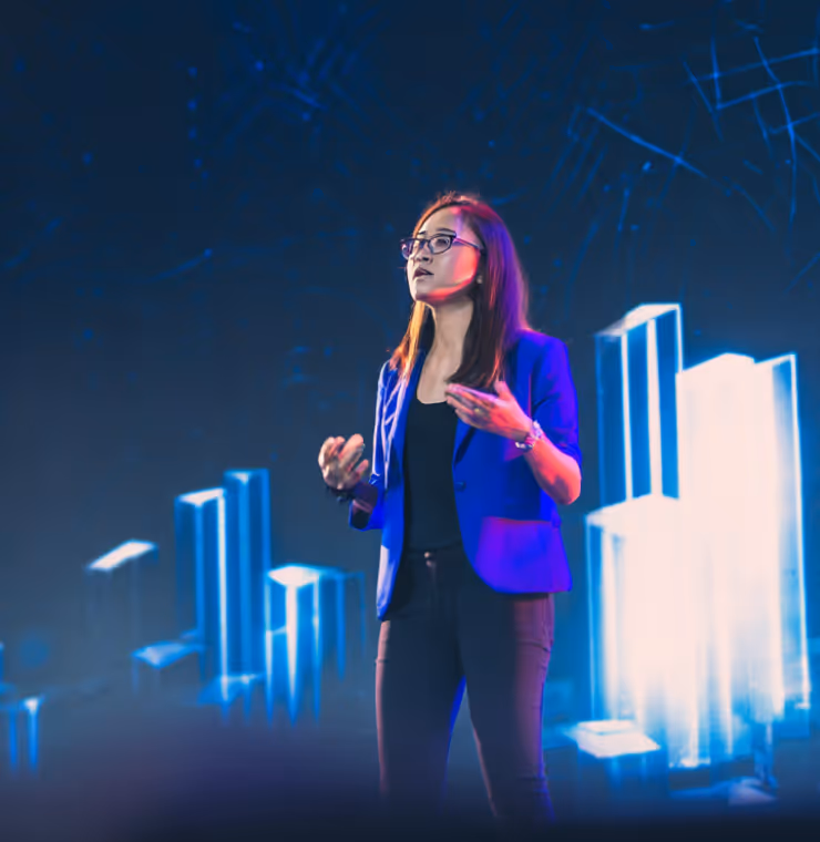 Woman in glasses and blue blazer giving a presentation with illuminated bar graphs in the background.