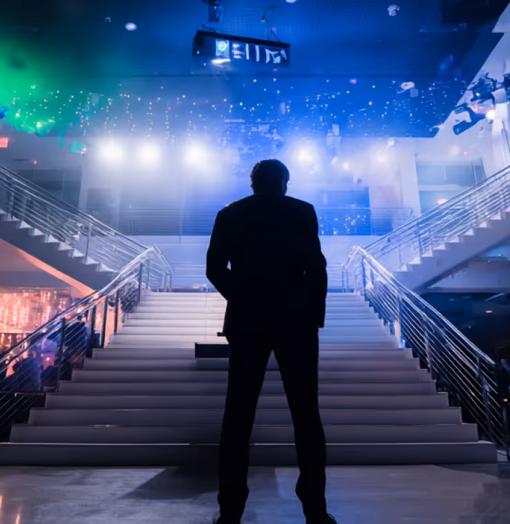 Silhouette of a man in a suit standing at the bottom of a wide staircase with bright lights and colorful reflections in a modern interior.