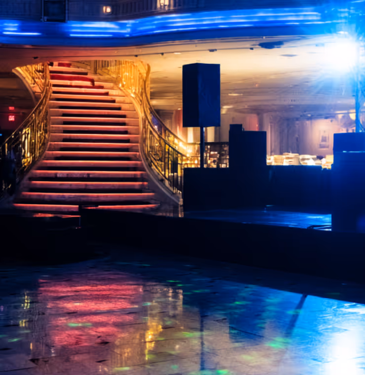 Elegant staircase with golden railings illuminated by colorful lights reflected on a glossy floor in a dimly lit venue.