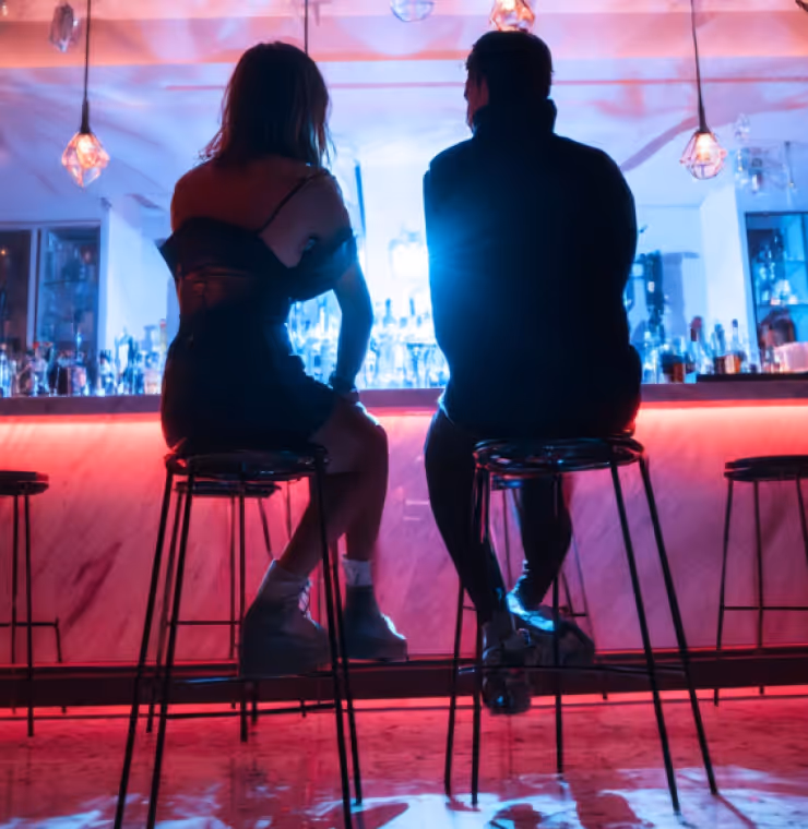 Silhouettes of a woman and a man sitting on bar stools at a backlit bar with colorful lighting and hanging pendant lamps.