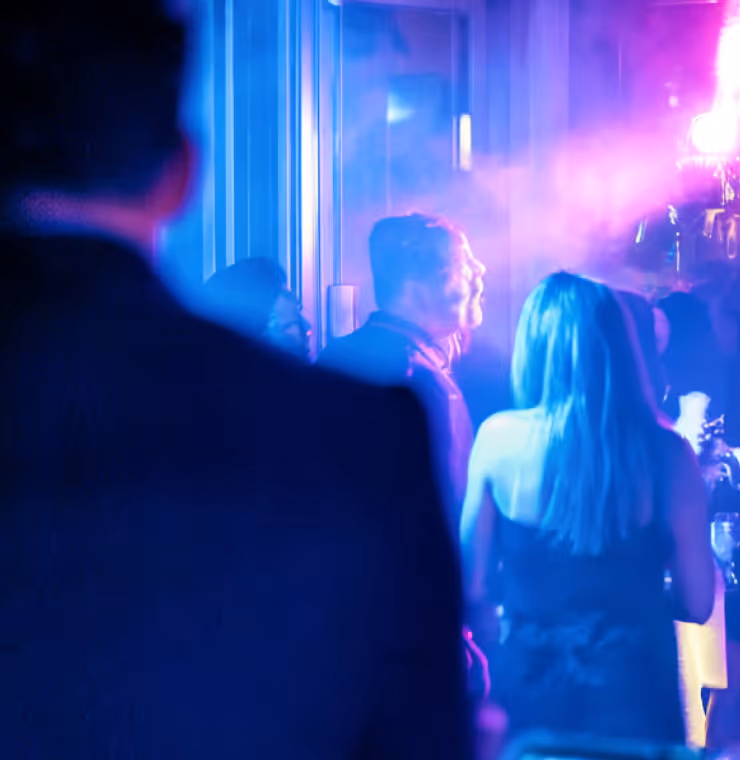 Group of people socializing in a dimly lit, blue and purple neon environment, possibly a nightclub or party.