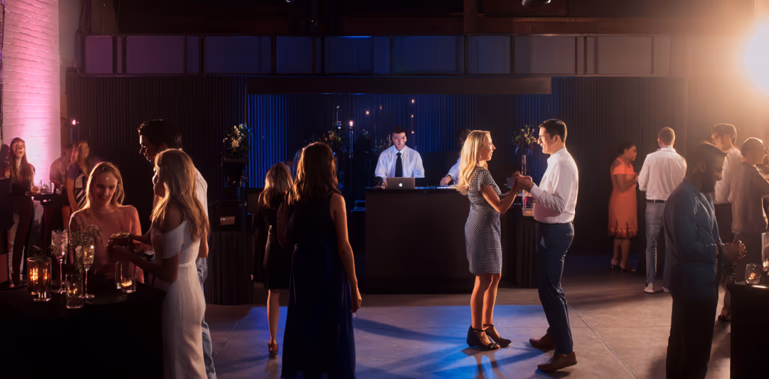 People socializing and dancing at an indoor party with a DJ booth and ambient lighting.