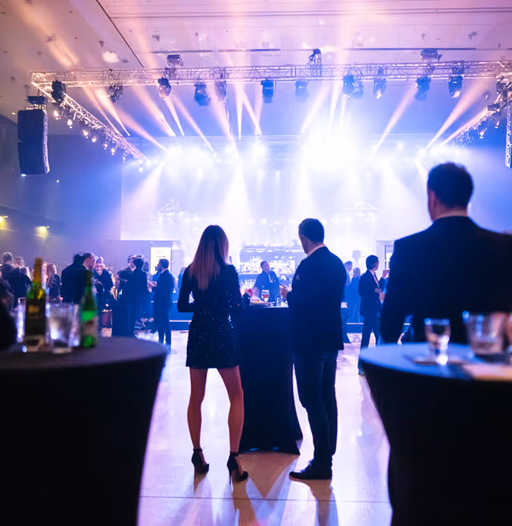 People socializing at a dimly lit event venue with bright stage lights and high tables.