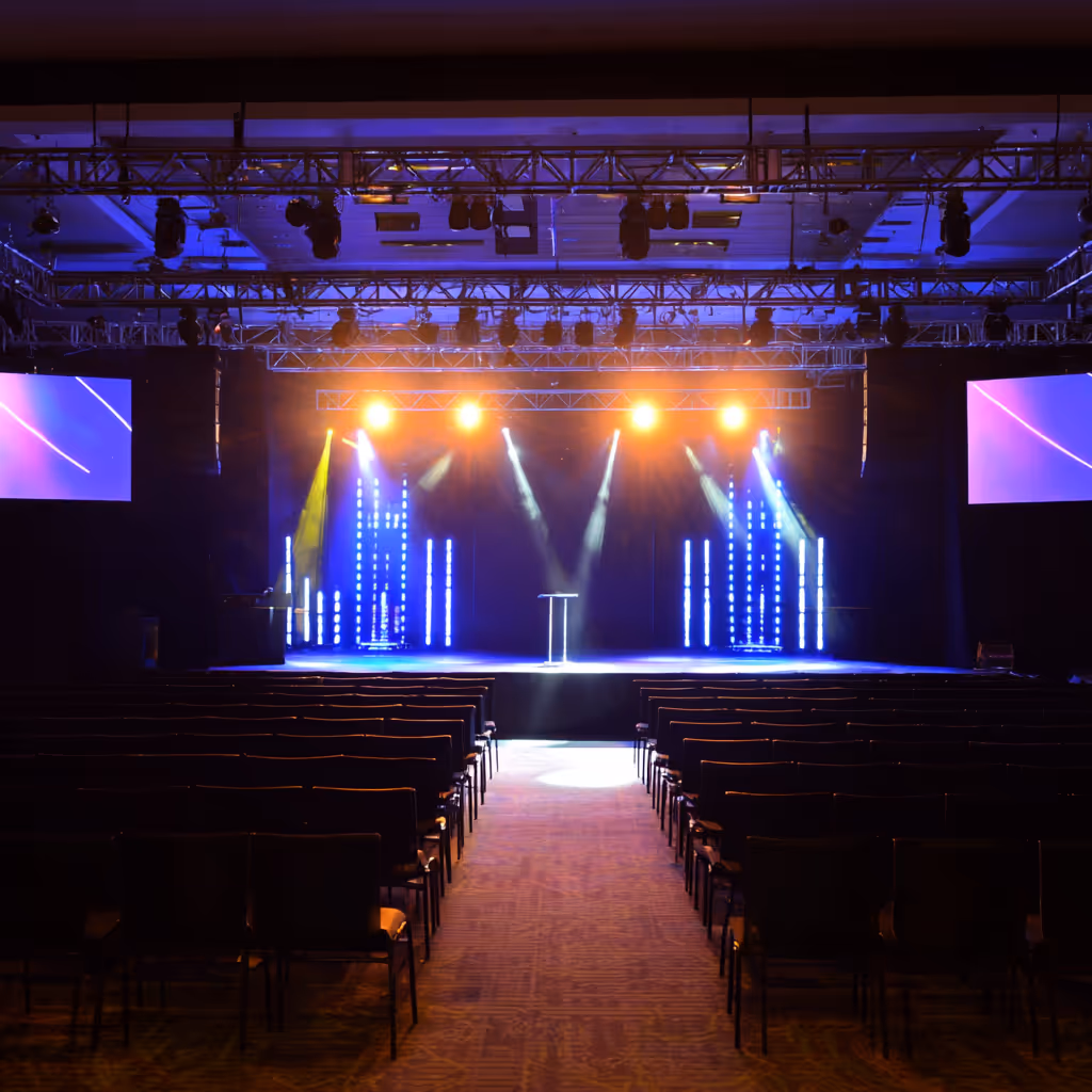 Empty auditorium with rows of chairs facing a stage illuminated by bright yellow and blue lights, with two large screens on either side.