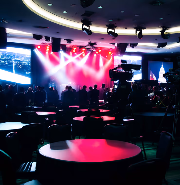 Dimly lit conference room with round tables and chairs, a crowd gathered near a stage with red lighting and large screens displaying cityscape and Capitol building images.