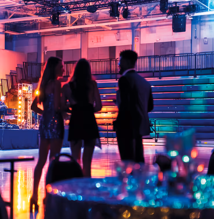 Three people in formal attire standing and talking in a colorful, dimly lit event space with tiered seating in the background.