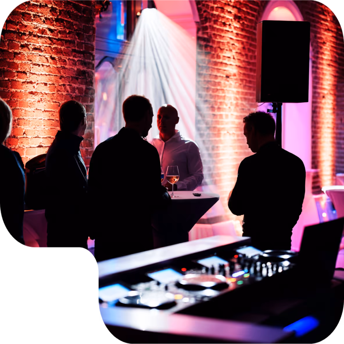 Silhouettes of people standing near a table with drinks in a dimly lit venue with colorful stage lights and a DJ setup in the foreground.