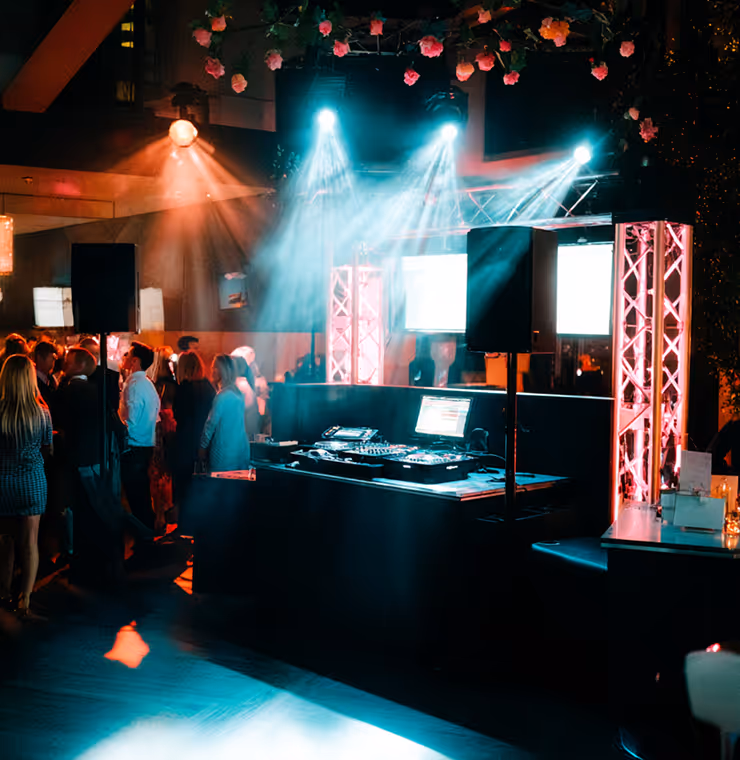 DJ booth with laptop and mixer illuminated by blue and pink stage lights at a crowded indoor event.