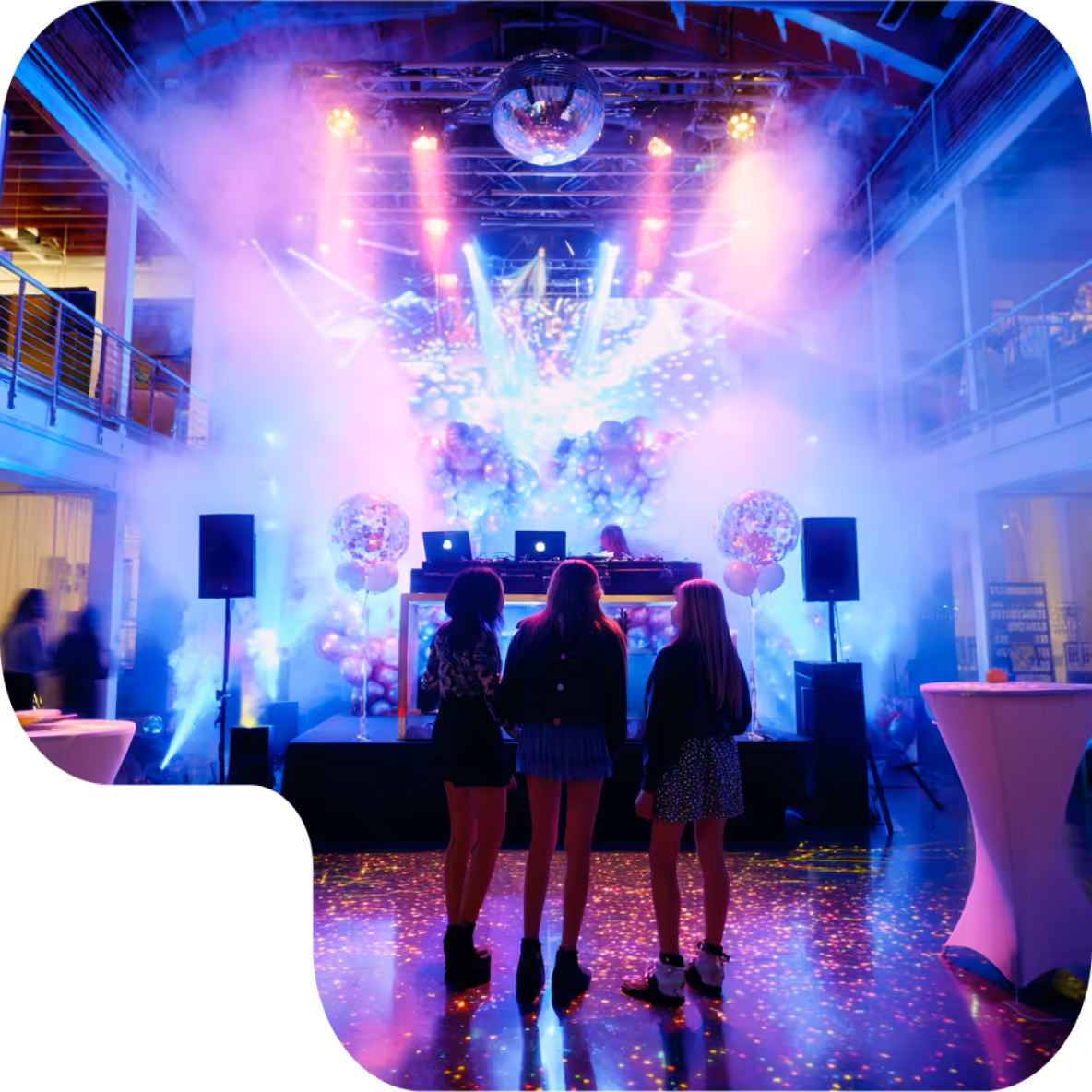 Three women standing in front of a DJ booth with colorful lights, fog, and balloons at an indoor party.