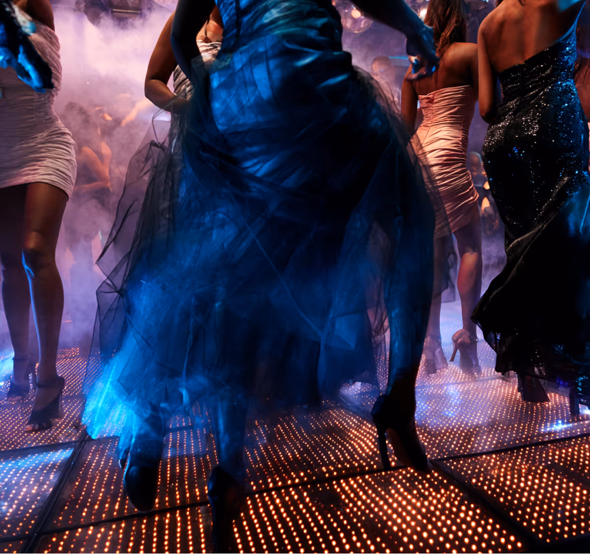 People wearing elegant dresses dancing on a lit-up floor with smoke effects in a club.