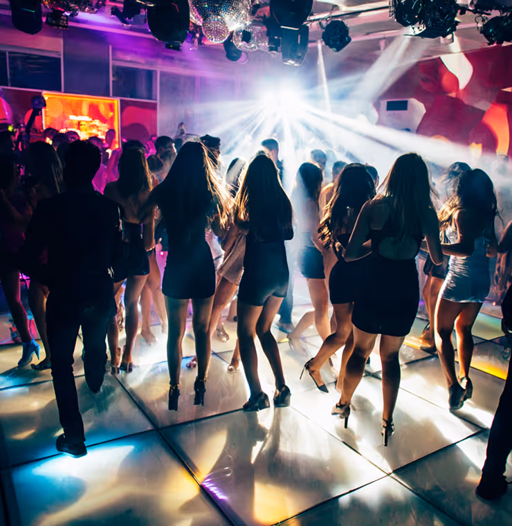 Group of people dancing on a brightly lit dance floor with colorful lights and fog effects in a nightclub.