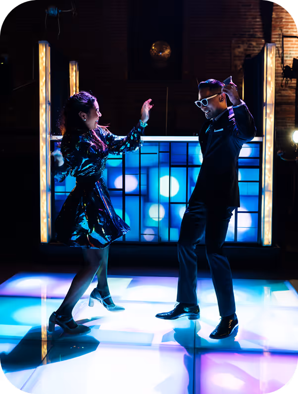 A man in a suit and sunglasses and a woman in a shiny dress dancing on a lit-up dance floor with colorful lights.