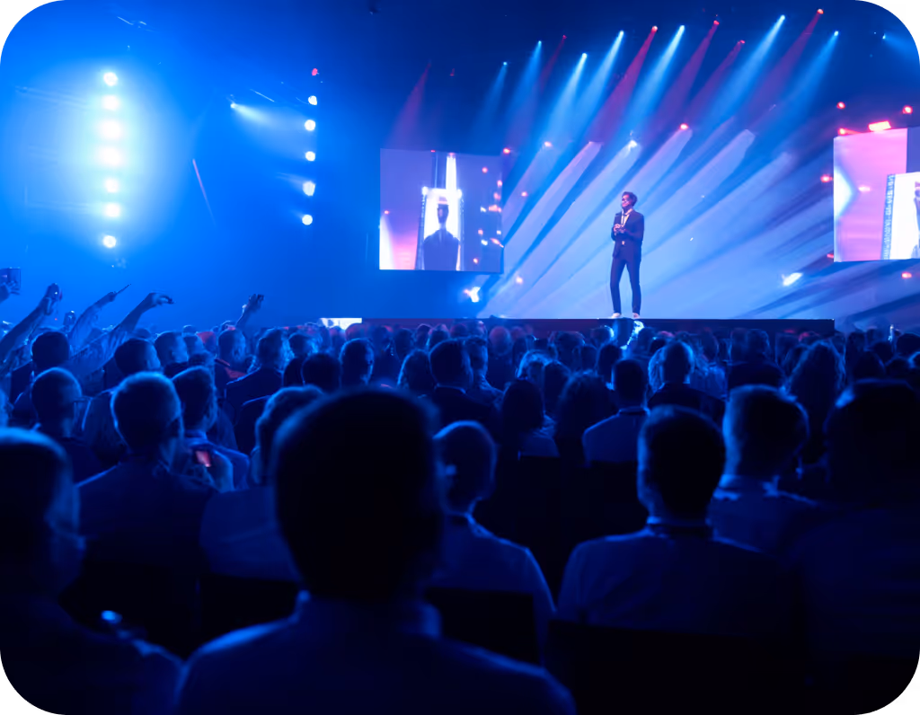 Speaker on stage in front of a large audience with blue and purple stage lighting and two large screens.
