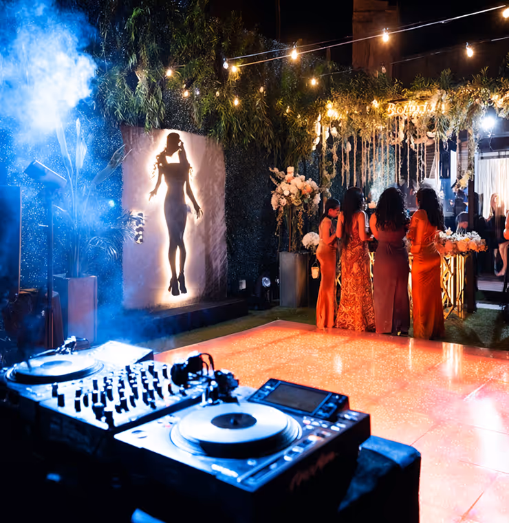 DJ turntables in foreground at night party with four women in elegant dresses near flower-decorated bar and illuminated silhouette backdrop.