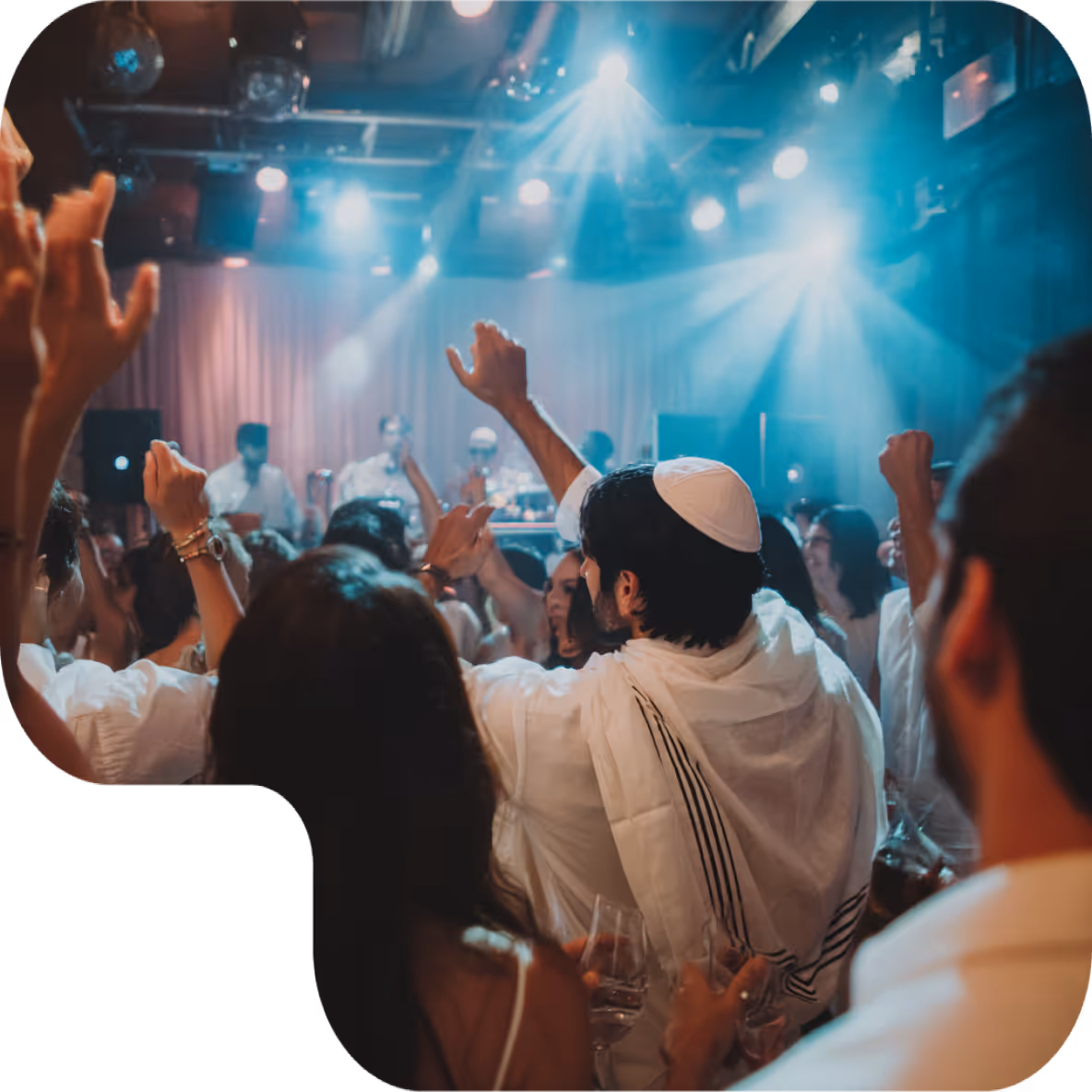 Crowd at a lively event with raised hands, a man wearing a kippah and tallit in foreground, illuminated by stage lights.