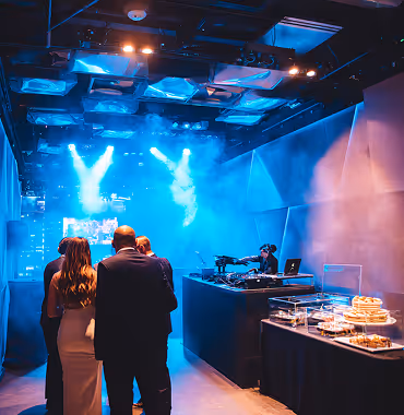Group of people standing near a DJ booth with colorful stage lights and fog in an indoor event space.