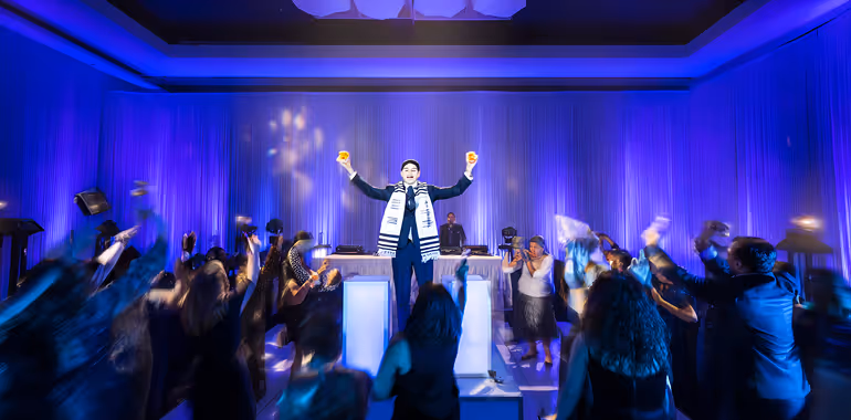 Man in formal attire holding up two drinks on a stage while people dance below in a blue-lit event space.