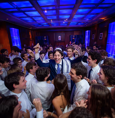 A young man wearing a tallit is celebrating joyfully surrounded by a crowd of people in a blue-lit room.