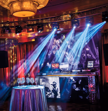 DJ setup with turntables and mixer under blue stage lights in a dimly lit room with red curtains and a table with empty wine glasses.