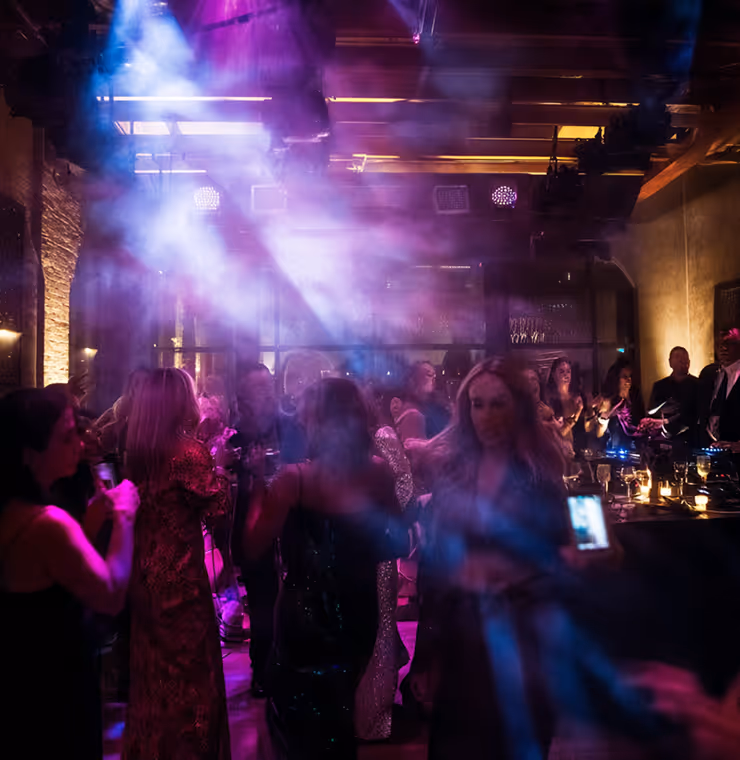 Dimly lit nightclub with people dancing and socializing under purple and blue stage lights and smoke effects.