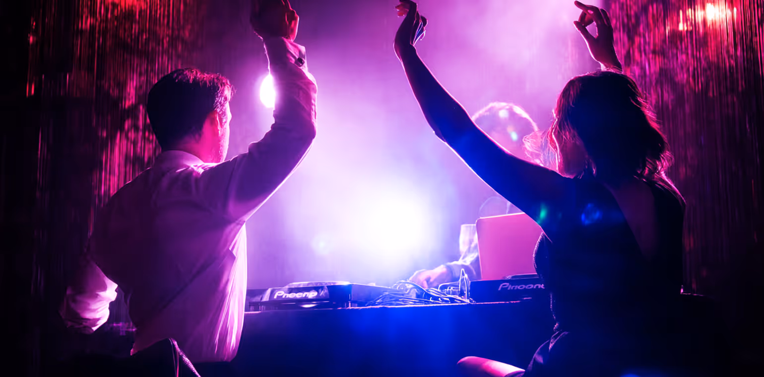 Two people dancing with raised arms near a DJ booth illuminated by vibrant purple and blue lights.