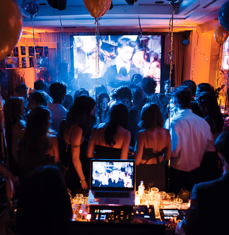 Crowd of people dancing at a dimly lit party with balloons and a large screen displaying a black-and-white video in the background.