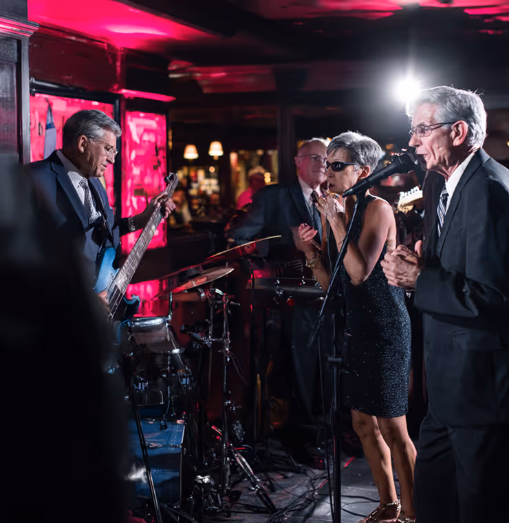 A senior band performing live on stage with a male guitarist, a female singer in sunglasses, and two male vocalists in suits under pink lighting.
