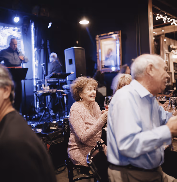 Elderly woman smiling and holding a glass of wine at a lively indoor social gathering with live music.