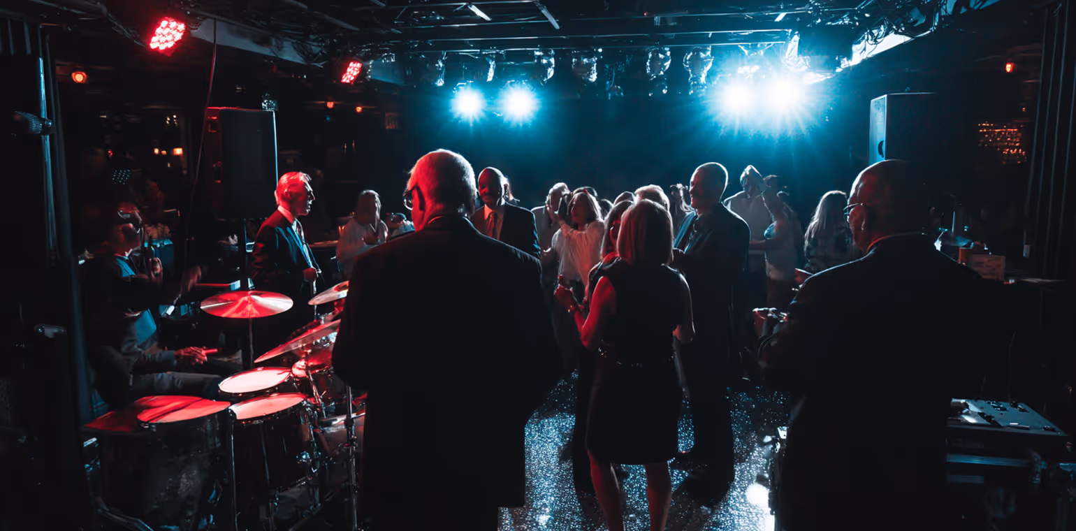 People dancing and socializing in a dimly lit club with a drummer playing on stage under blue and red lights.