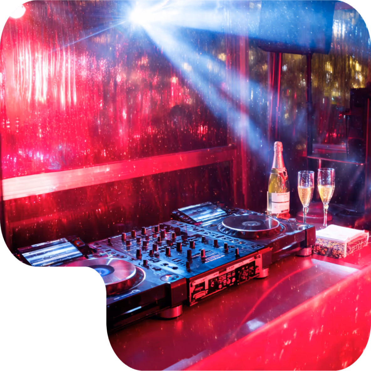 DJ mixer and turntables on a table with a bottle of champagne and two filled glasses, illuminated by colorful party lights.