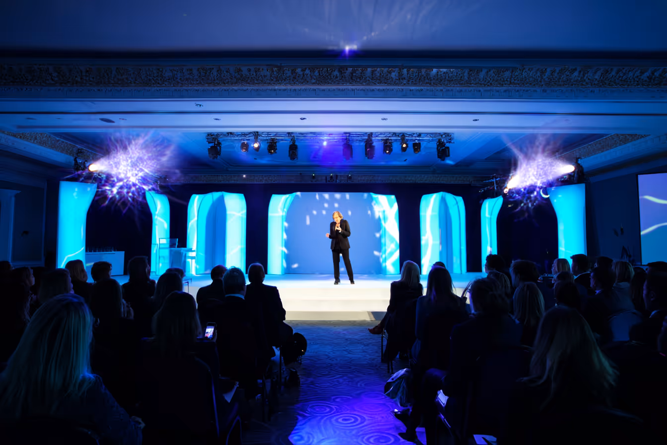 Speaker in black suit presenting on stage with blue illuminated backdrop and seated audience in a conference room.