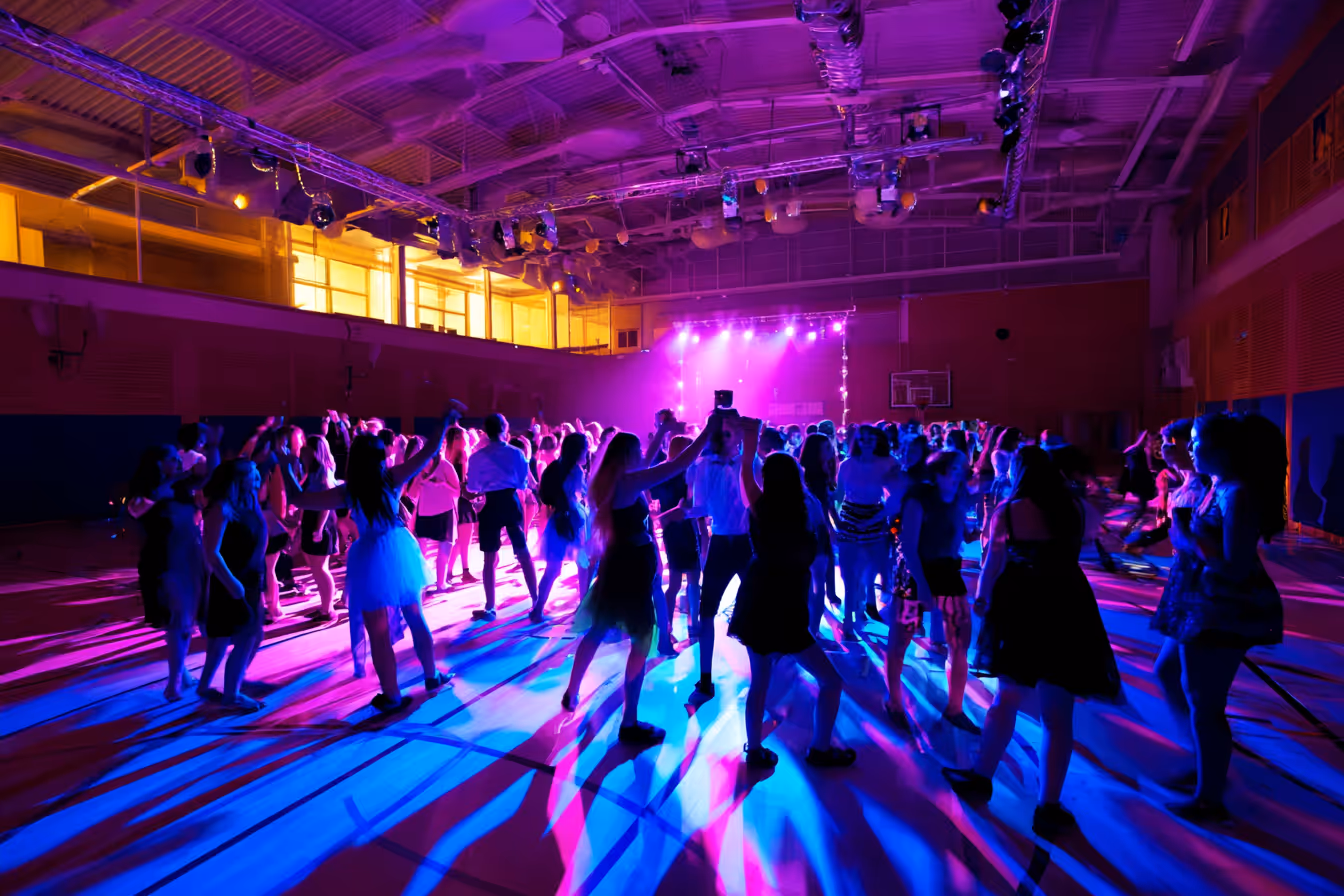 Group of people dancing in a gymnasium with vibrant purple and blue stage lighting.