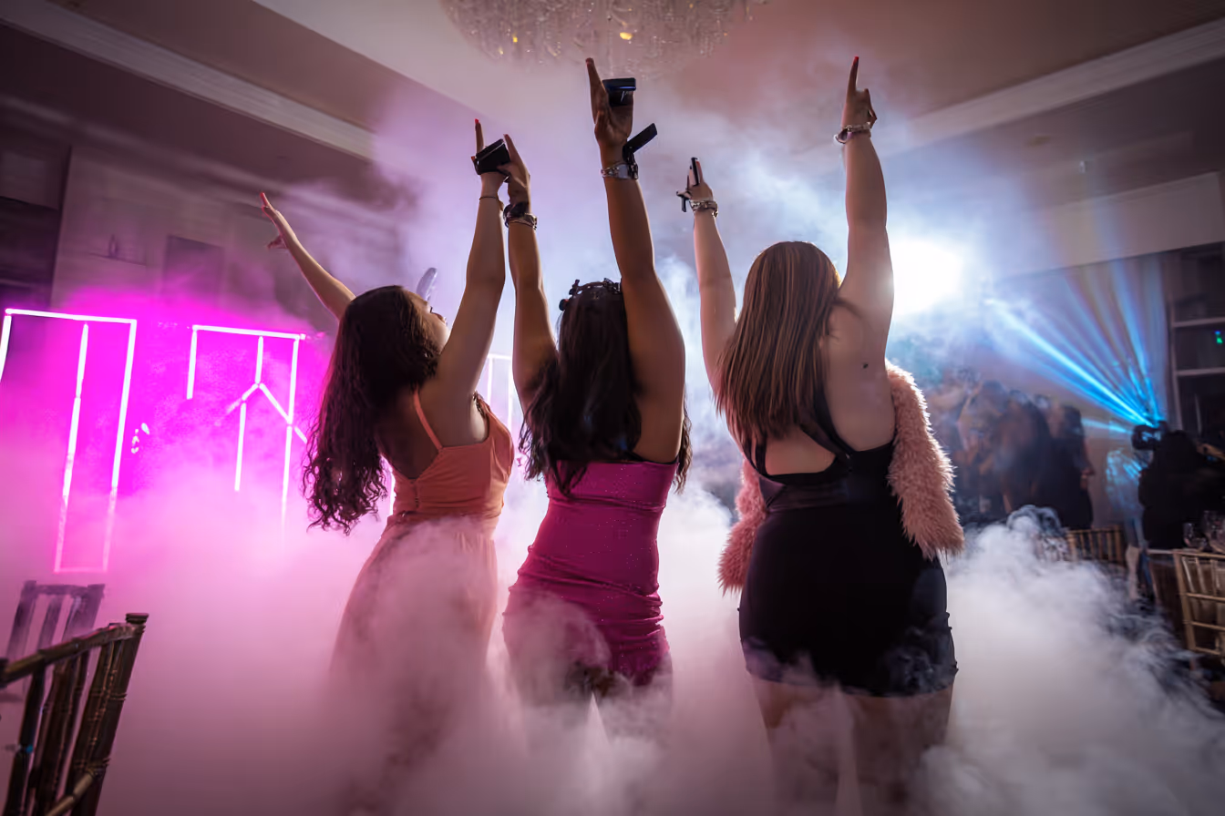 Three women in party dresses with their hands raised, surrounded by fog and colorful lights at a nightclub or celebration.
