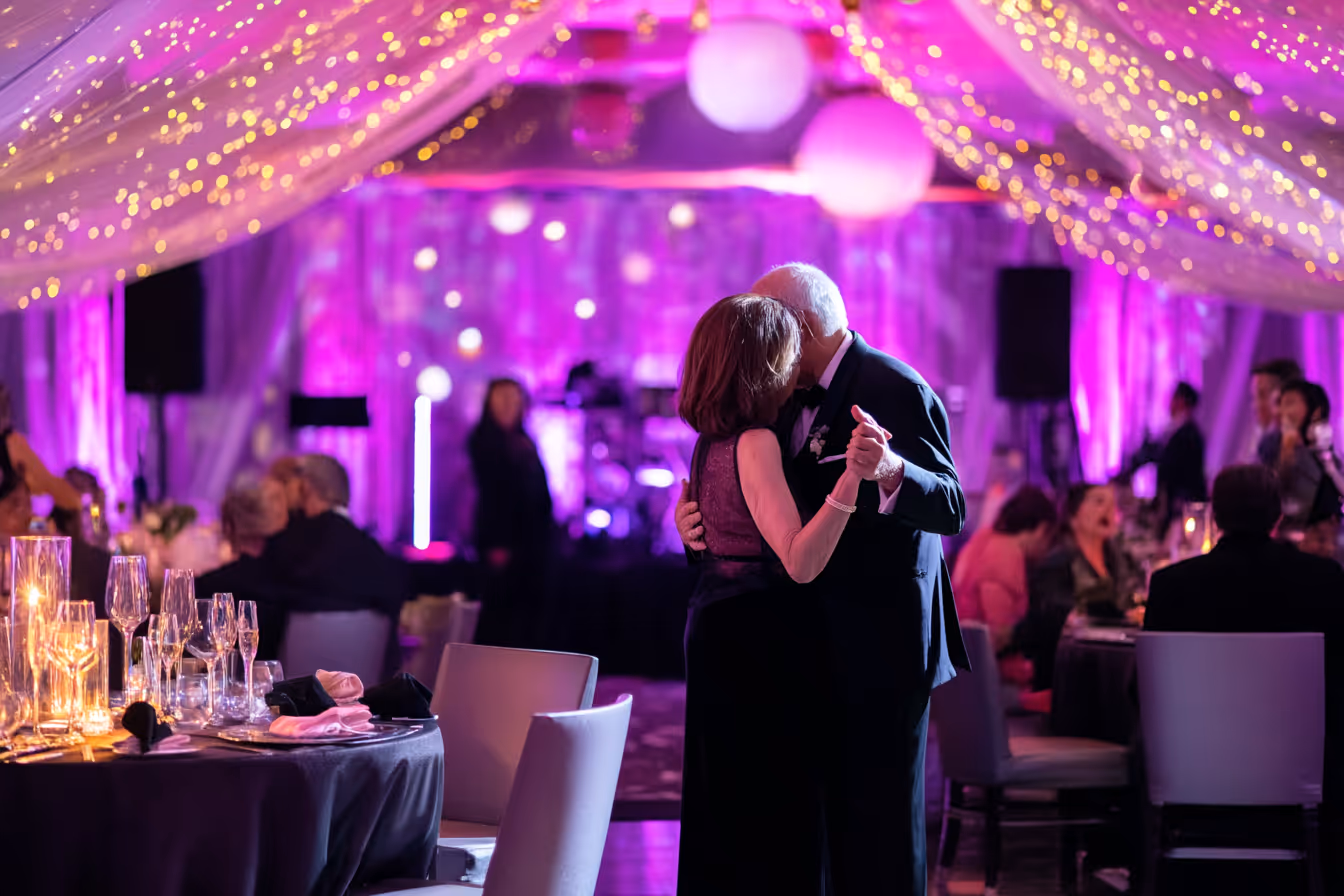 Elderly couple dancing closely at a formal event with purple lighting and string lights overhead.