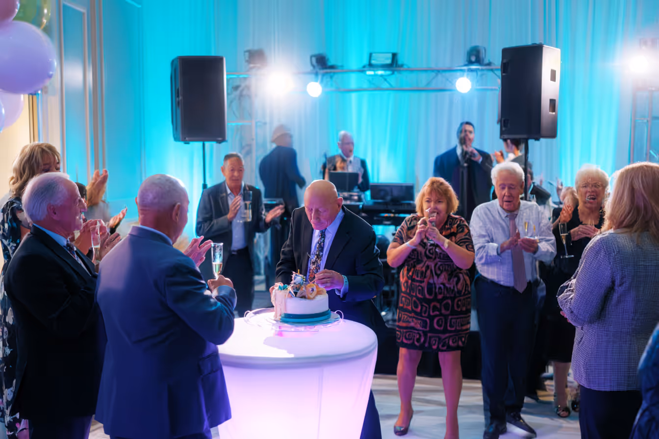 Elderly man cutting a cake at a party while guests around him clap and hold drinks, with musicians and blue lighting in the background.