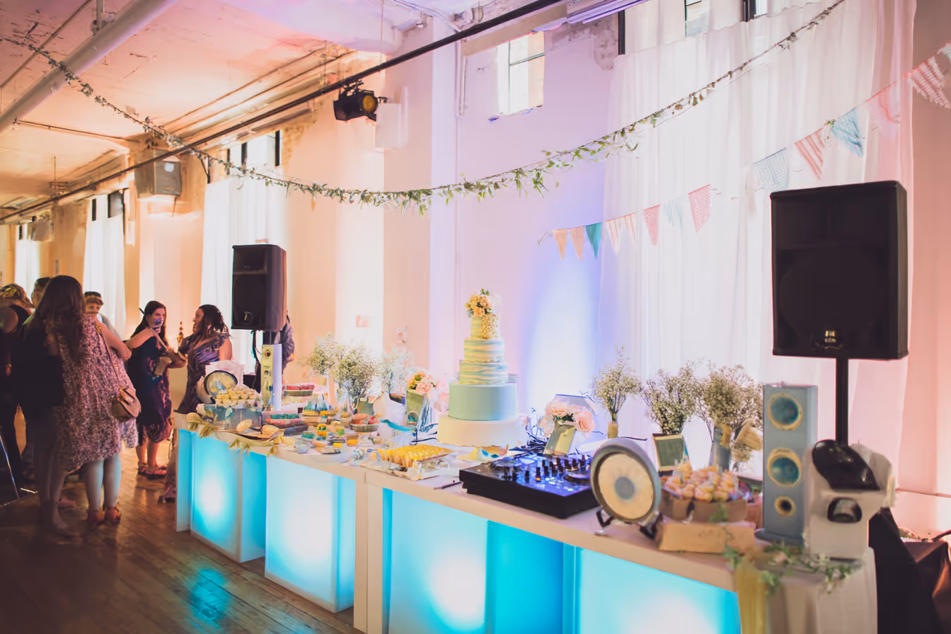 Decorated dessert table with a tiered cake, pastries, flowers, and DJ equipment lit with blue lights at an indoor party with guests nearby.