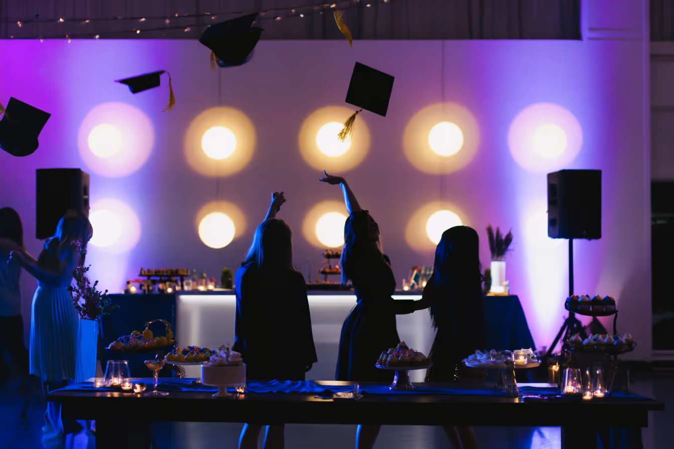 Silhouettes of graduates throwing black mortarboards in the air at a dimly lit party with purple and yellow lights.