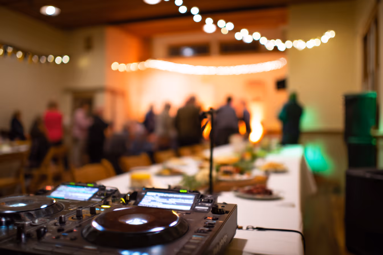 DJ turntables in focus with a blurred background of people mingling in a warmly lit room decorated with string lights.