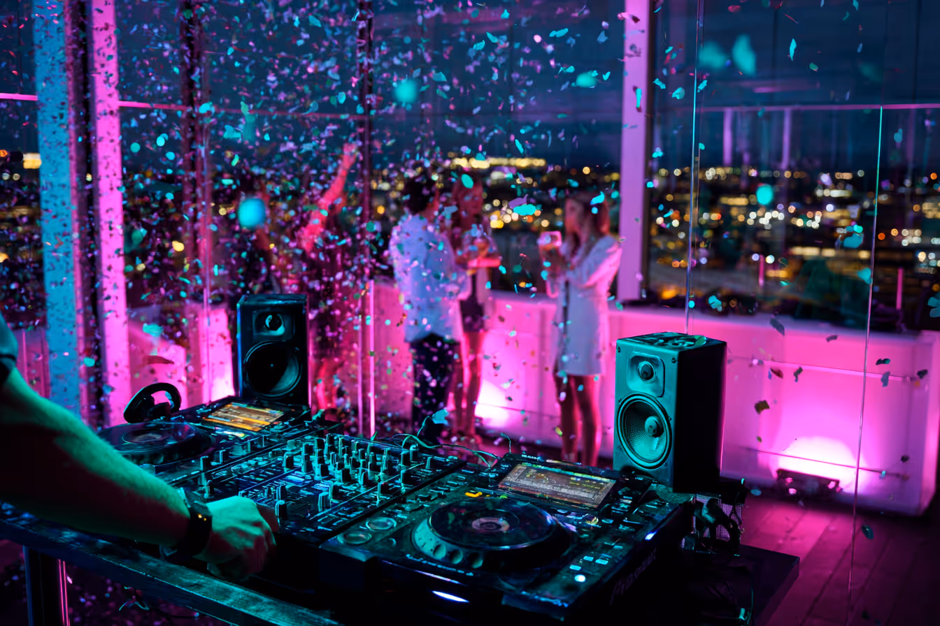DJ playing music on a mixer at a rooftop party with confetti falling and people socializing in the background.