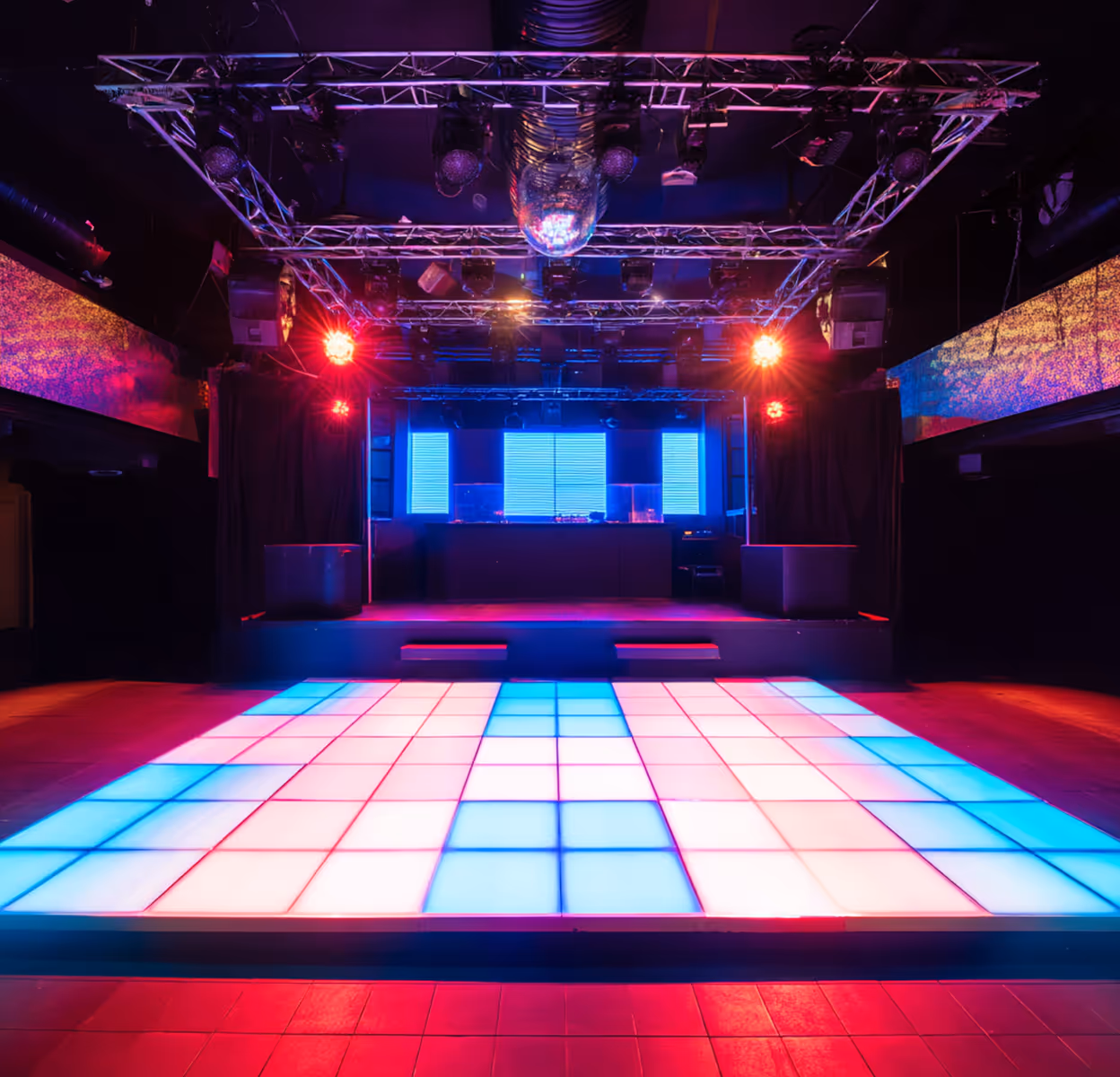 Empty nightclub dance floor illuminated with blue and pink LED lights in front of a DJ stage with bright screens and lighting equipment.